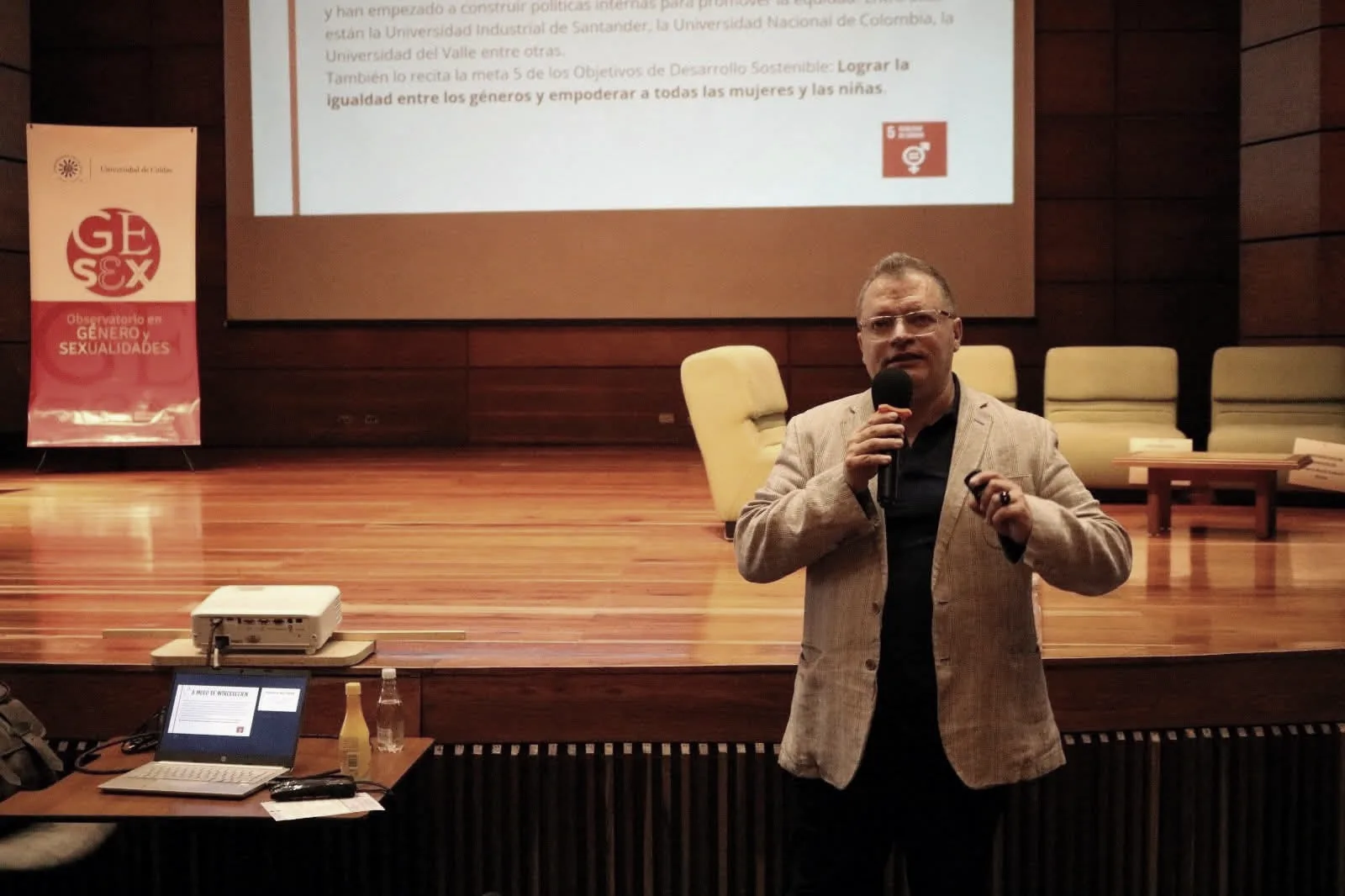 José Fernando Vasco Alzate habla con micrófono en un auditorio. Detrás, una diapositiva sobre el Objetivo de Desarrollo Sostenible 5 y el pendón del Observatorio en Género y Sexualidades de la Universidad de Caldas.